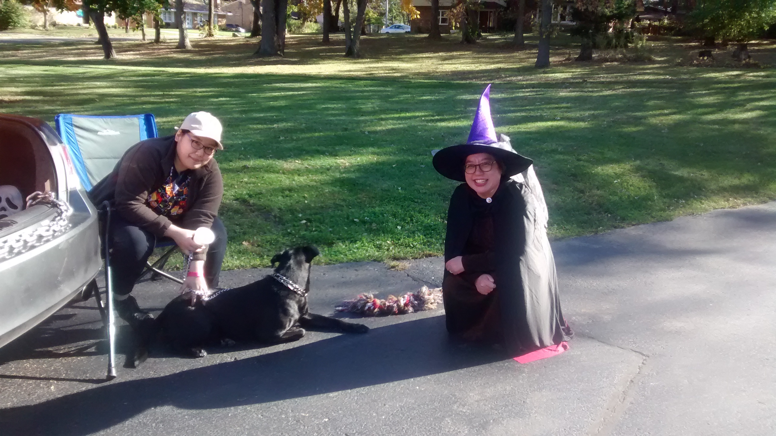 A pastor dressed up as a witch crouching next to a seated person and dog.
